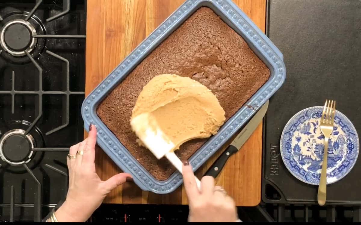 A person spreads beige frosting over a rectangular chocolate cake using a spatula. The cake is in a blue baking dish on a wooden cutting board. Nearby, there is a knife and a decorated plate with two forks.