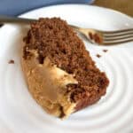 A slice of chocolate cake with peanut butter frosting sits on a white plate next to a fork. The cake appears moist and is partially eaten, with crumbs scattered on the plate.