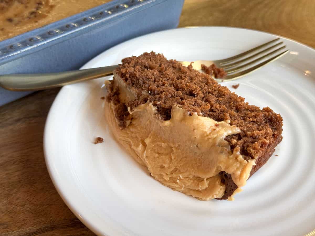 A slice of chocolate cake with caramel frosting on a white plate, with a fork beside it. The background shows the rest of the cake in a blue baking pan on a wooden surface.