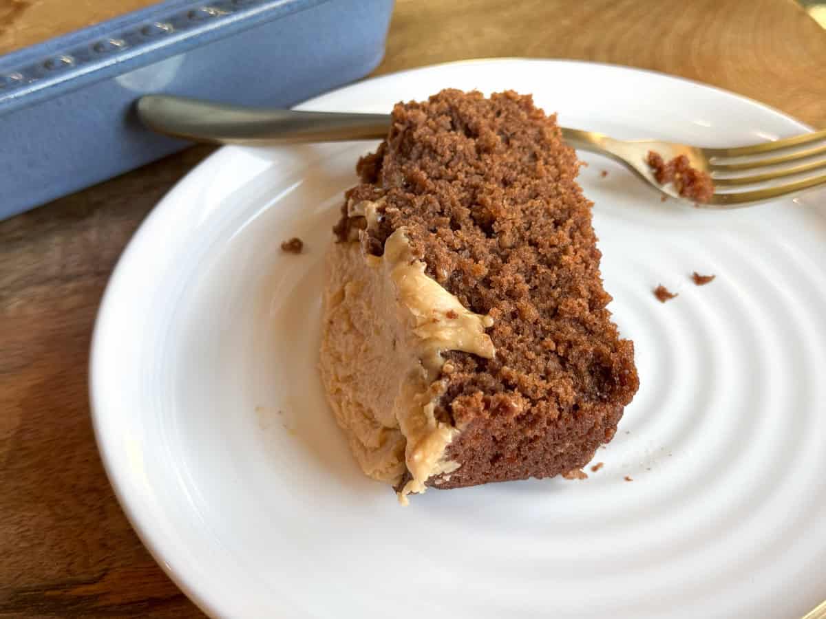 A slice of chocolate cake with frosting on a white plate, partially eaten. A silver fork rests on the edge of the plate. A blue baking dish is visible in the background on a wooden table.