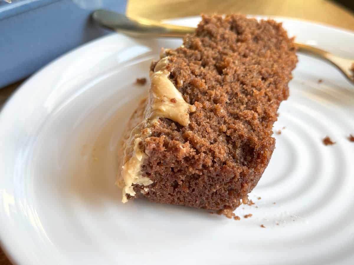 A slice of chocolate cake with creamy frosting on a white plate. A silver fork is placed beside the cake.
