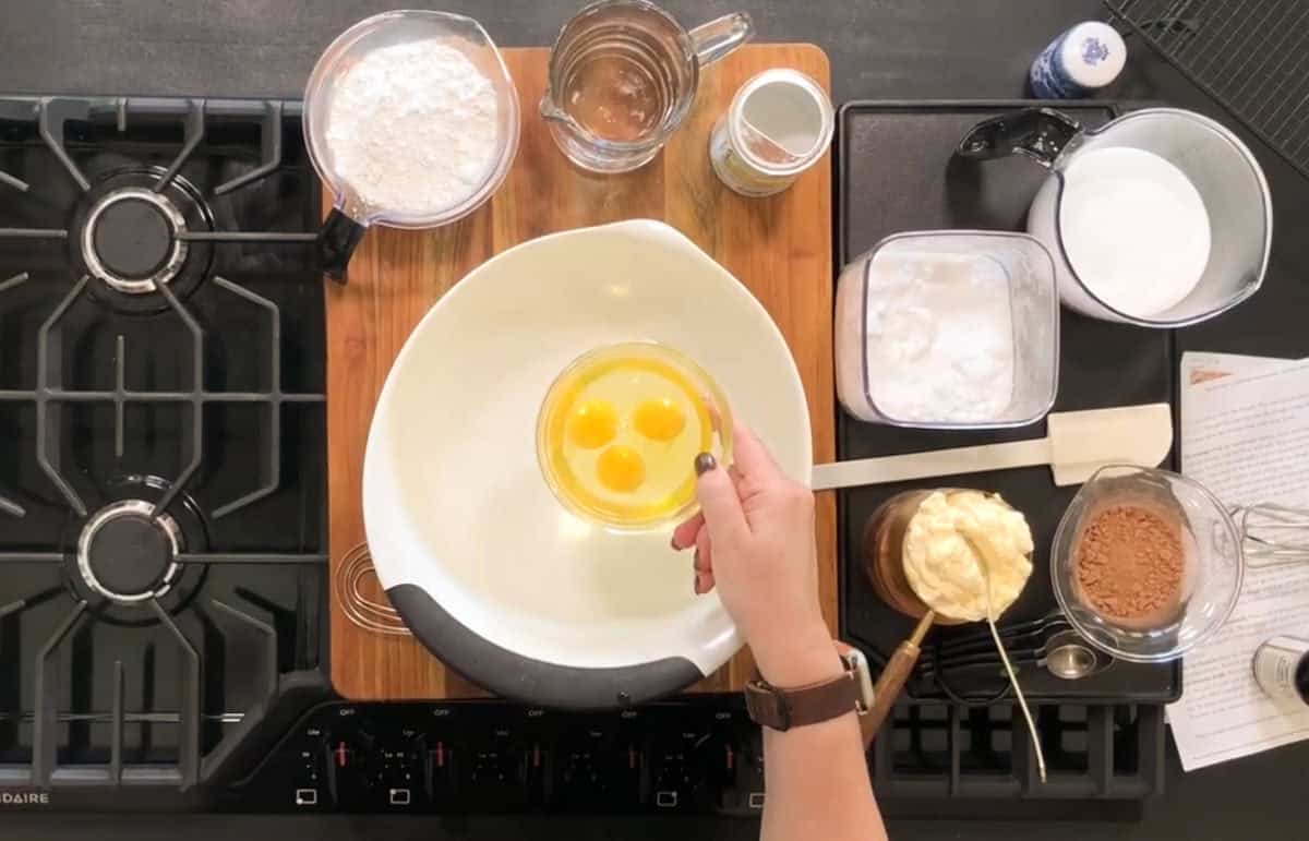A person holds a bowl with three raw eggs above a mixing bowl. Surrounding ingredients include flour, sugar, cocoa powder, butter, and a measuring cup with water. The setup is on a kitchen countertop with a stove on the left.