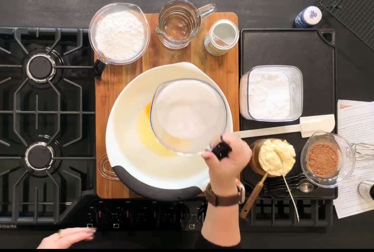 A person is preparing ingredients for baking on a stovetop. A mixing bowl and various ingredients, including flour, cocoa powder, and butter, are arranged on a wooden board. A hand is holding a measuring cup over the bowl.