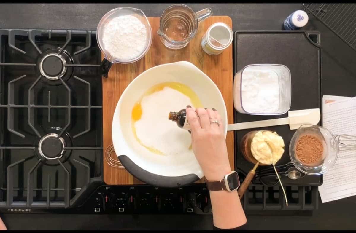 Top view of a person adding vanilla extract to a mixing bowl on a stovetop. The bowl contains eggs and other baking ingredients. Surrounding the bowl are flour, baking soda, a recipe sheet, butter, cocoa, and a mixer. A whisk and measuring cup are also visible.