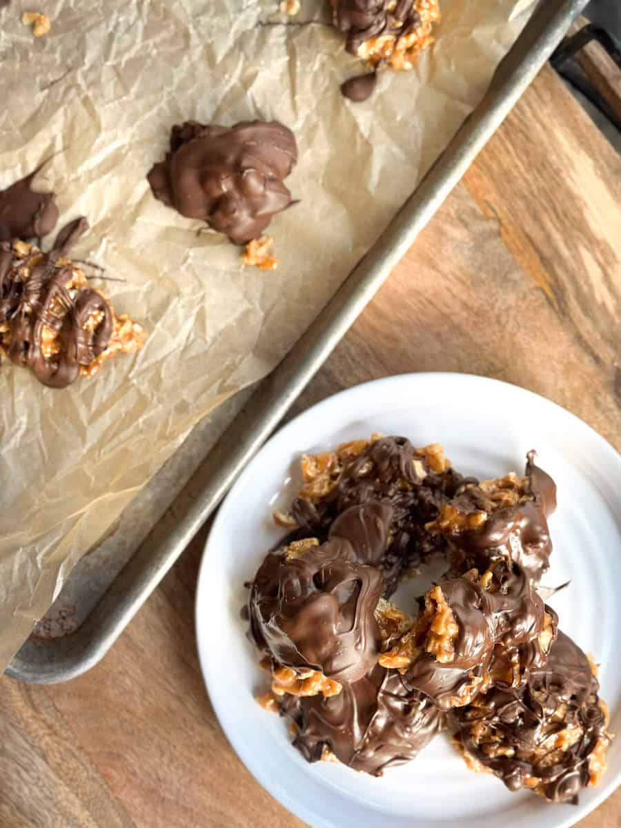 A baking tray with parchment paper holds chocolate-covered clusters. Beside it, a white plate displays more of the same chocolate clusters, with a wooden surface underneath. The clusters have a rough, irregular shape.