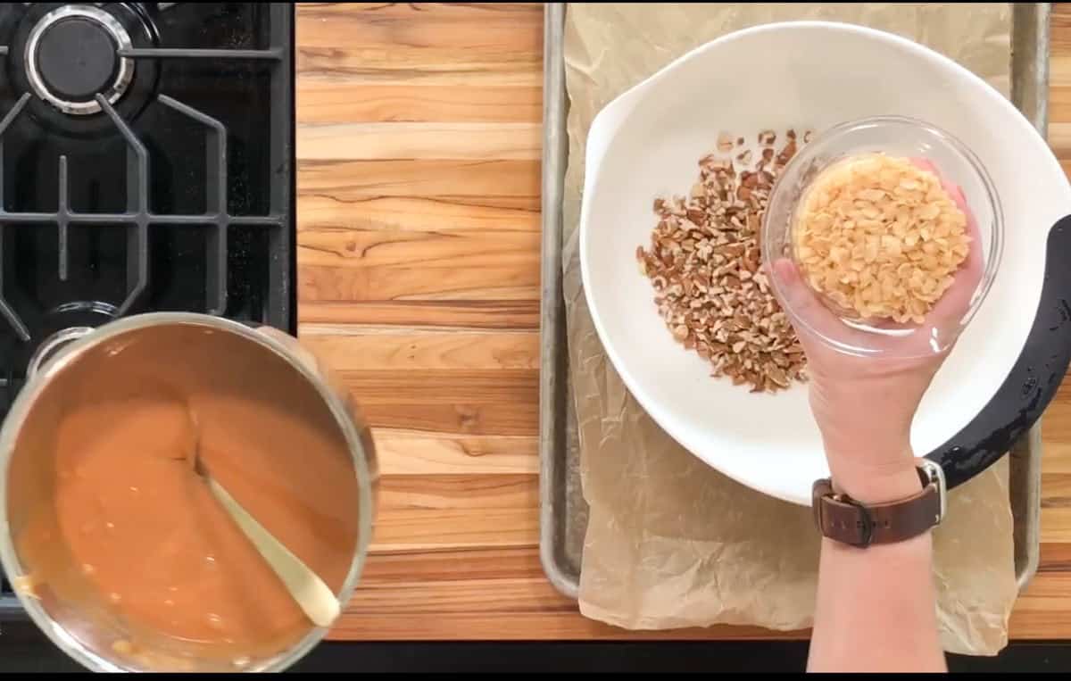 Overhead view of a person holding a small bowl of sliced almonds above a larger bowl containing chopped nuts. A pot with a caramel-colored mixture and a stove are visible on a wooden countertop.