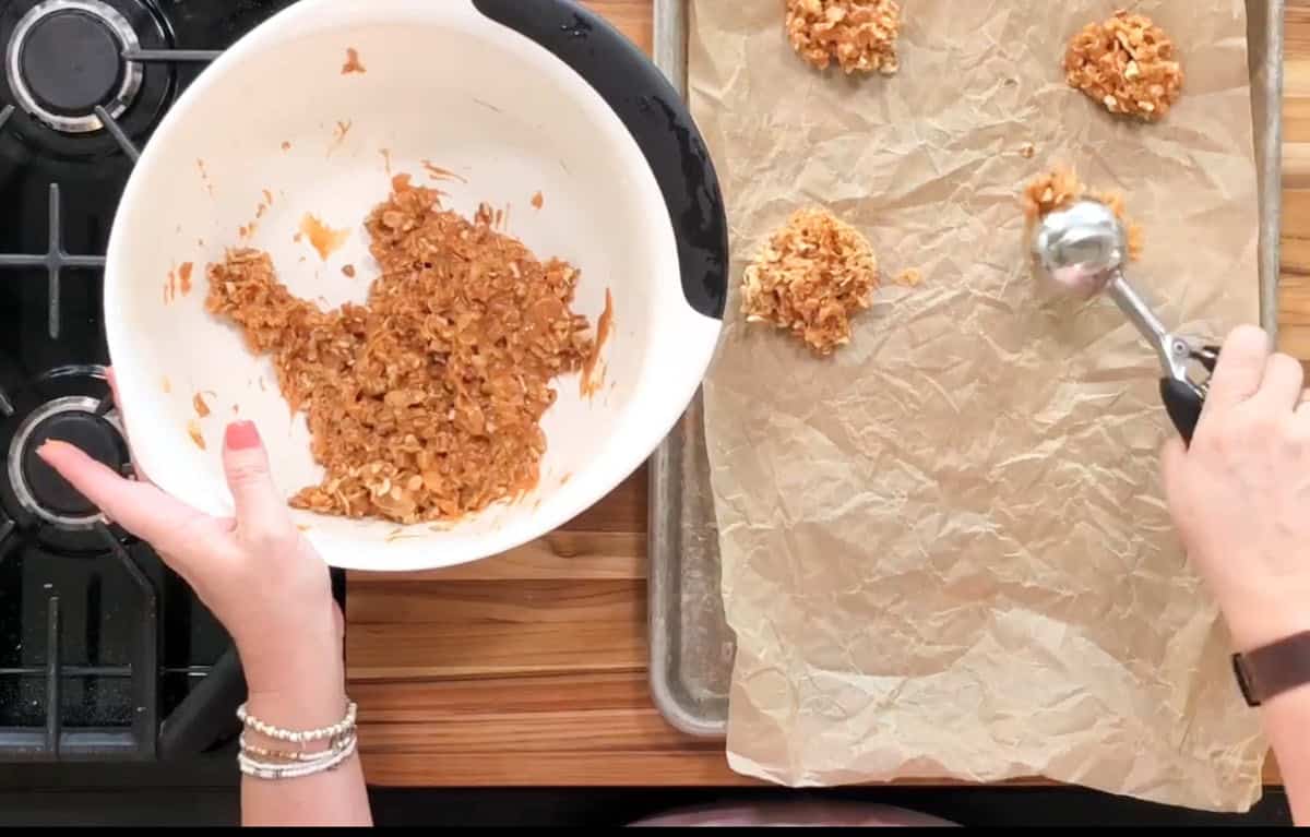 A person is scooping cookie dough from a mixing bowl onto a parchment-lined baking sheet with a cookie scoop. The dough appears to contain oats. The baking sheet is placed on a wooden countertop next to a stove.