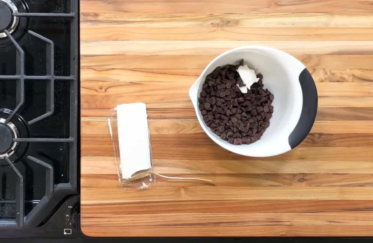 A white mixing bowl with chocolate chips and a piece of butter on a wooden countertop. Next to it, a wrapped stick of butter sits on the counter. A black stove is partially visible on the left.
