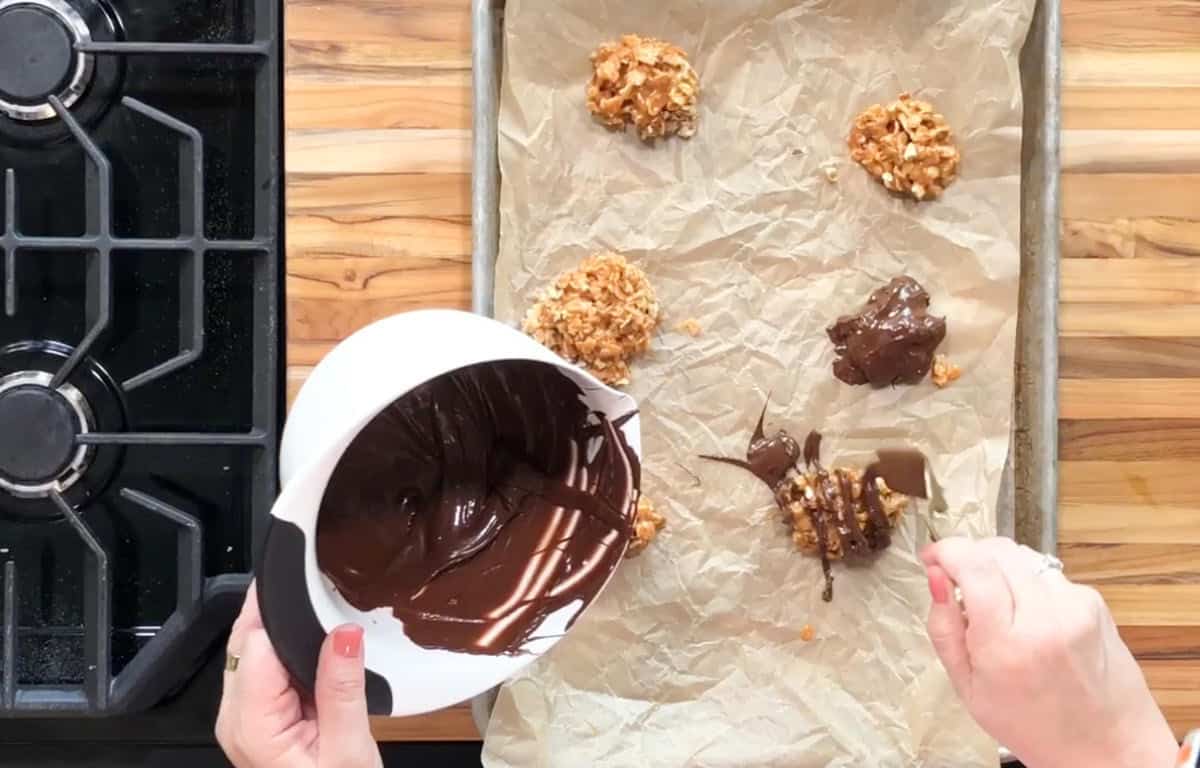 A person is spooning melted chocolate onto oat-based cookies on a parchment-lined baking sheet. The tray is on a wooden countertop next to a black stovetop.