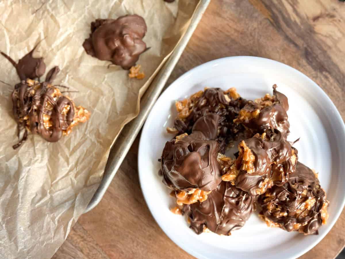 Chocolate-covered clusters are placed on a white plate. Nearby, a baking tray with parchment paper holds additional clusters. The clusters appear to contain nuts or cereal, drizzled or coated with chocolate, set on a wooden surface.