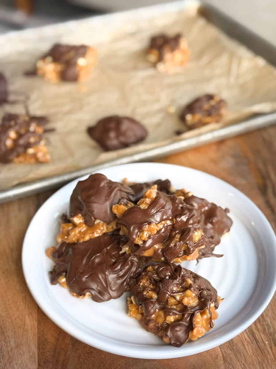 A white plate with several clusters of caramel corn covered in melted chocolate. In the background, a baking tray with more chocolate-coated caramel corn sits on parchment paper. The scene is set on a wooden surface.
