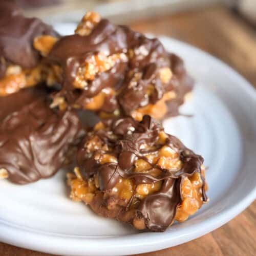 A close-up of caramel and chocolate-covered clusters on a white plate. The clusters appear to contain nuts and are drizzled with melted chocolate, creating a glossy texture. The plate is positioned on a wooden surface.