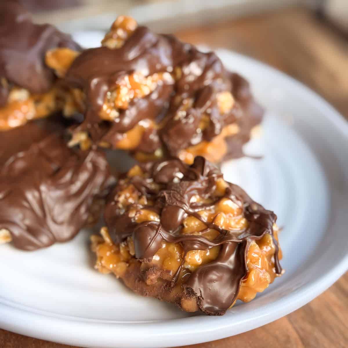 A close-up of caramel and chocolate-covered clusters on a white plate. The clusters appear to contain nuts and are drizzled with melted chocolate, creating a glossy texture. The plate is positioned on a wooden surface.