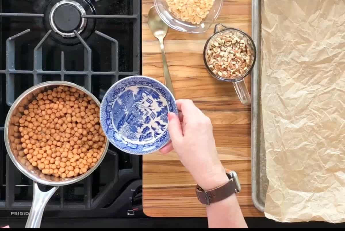 A person pours spices from a small blue and white bowl into a pot of chickpeas on a stove. Nearby are chopped nuts in a cup and a bowl, with a spoon and a baking tray lined with parchment paper on a wooden surface.