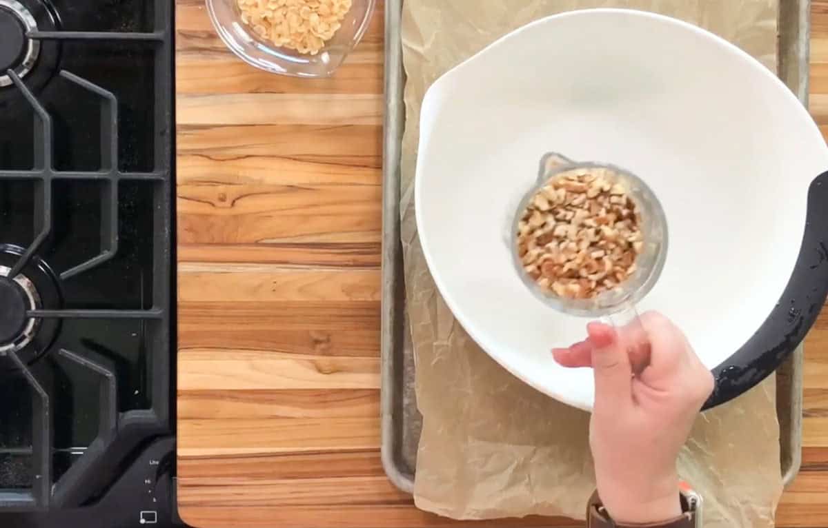 A person's hand holds a measuring cup filled with chopped nuts over a large white mixing bowl. The bowl sits on a baking tray lined with parchment paper. A small glass bowl with more nuts is nearby on a wooden countertop next to a stove.