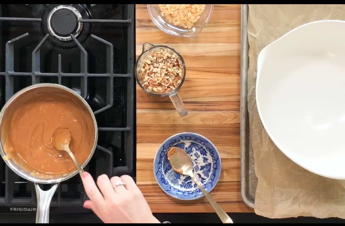 A stovetop scene with a saucepan containing caramel being stirred by a hand. Nearby are chopped nuts in a measuring cup and a blue-patterned plate with a spoon. A large white bowl and a lined baking sheet are on the wooden counter.