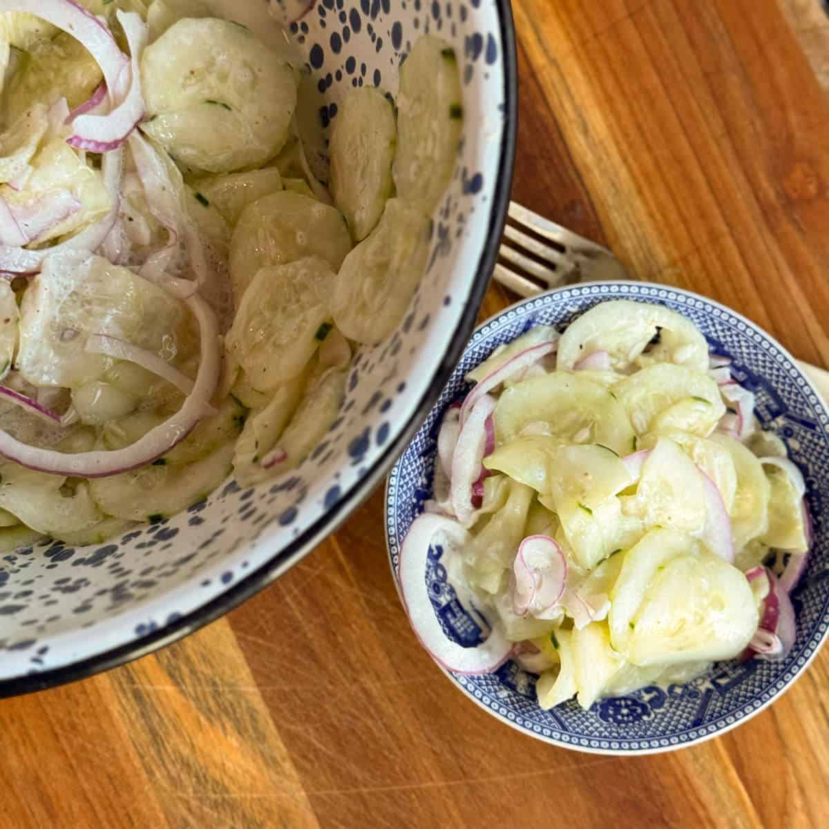 A large bowl and a smaller bowl, both containing sliced cucumber and red onion in a creamy dressing, are placed on a wooden surface. A fork lies next to the smaller bowl.