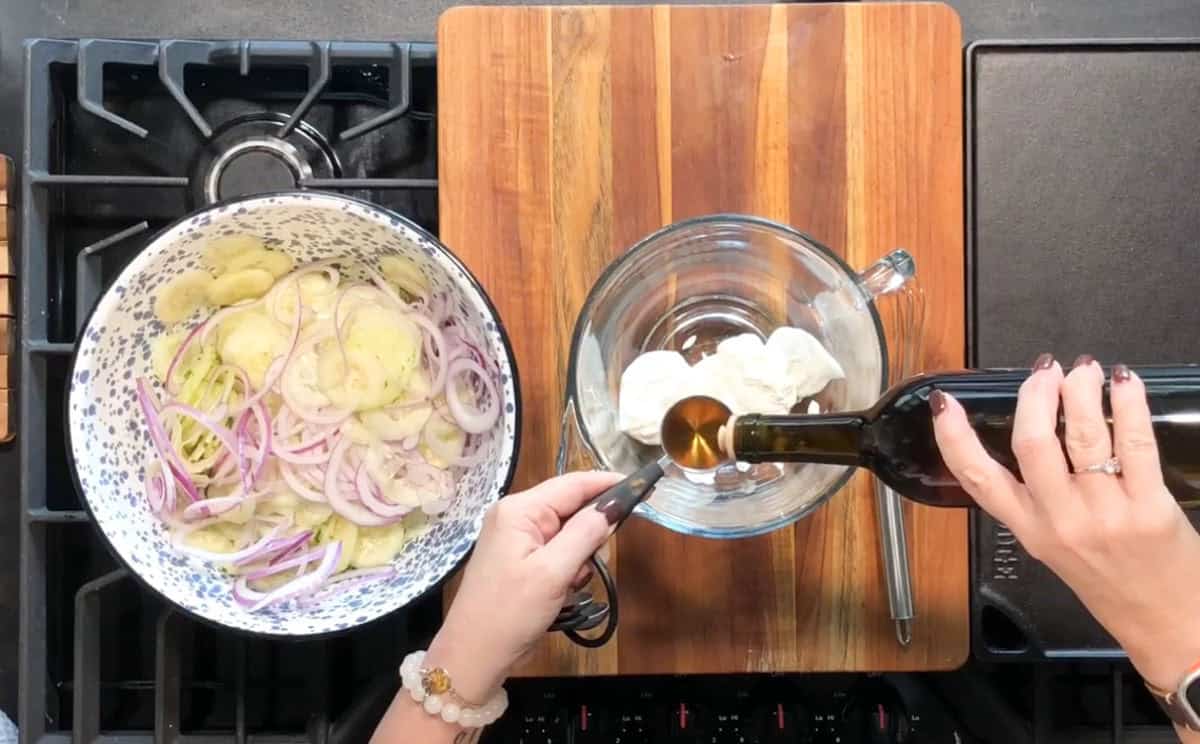 A person prepares a dressing by pouring olive oil into a glass bowl containing mayonnaise. Next to it, there is a large pot on a stove filled with sliced potatoes and onions. A wooden cutting board is placed between the pot and the bowl.