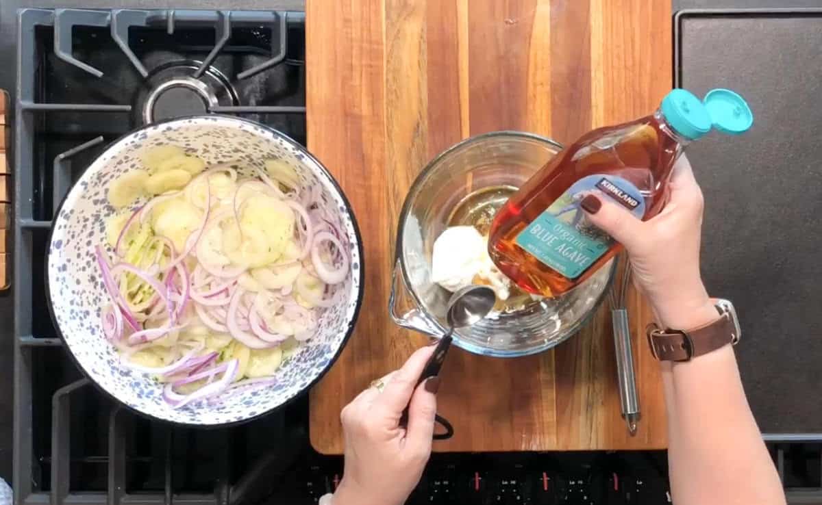 A person's hands are preparing a dish. On a wooden surface, there's a bowl with sliced onions and cucumbers. Another glass bowl holds white sauce being mixed with a spoon, and there's a bottle of apple cider vinegar nearby.