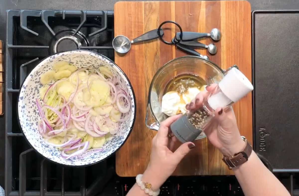 A person grinds pepper into a bowl containing mayonnaise and other ingredients. On the left, a large bowl holds sliced potatoes and red onions. Measuring spoons and a wooden cutting board are visible on the stovetop.