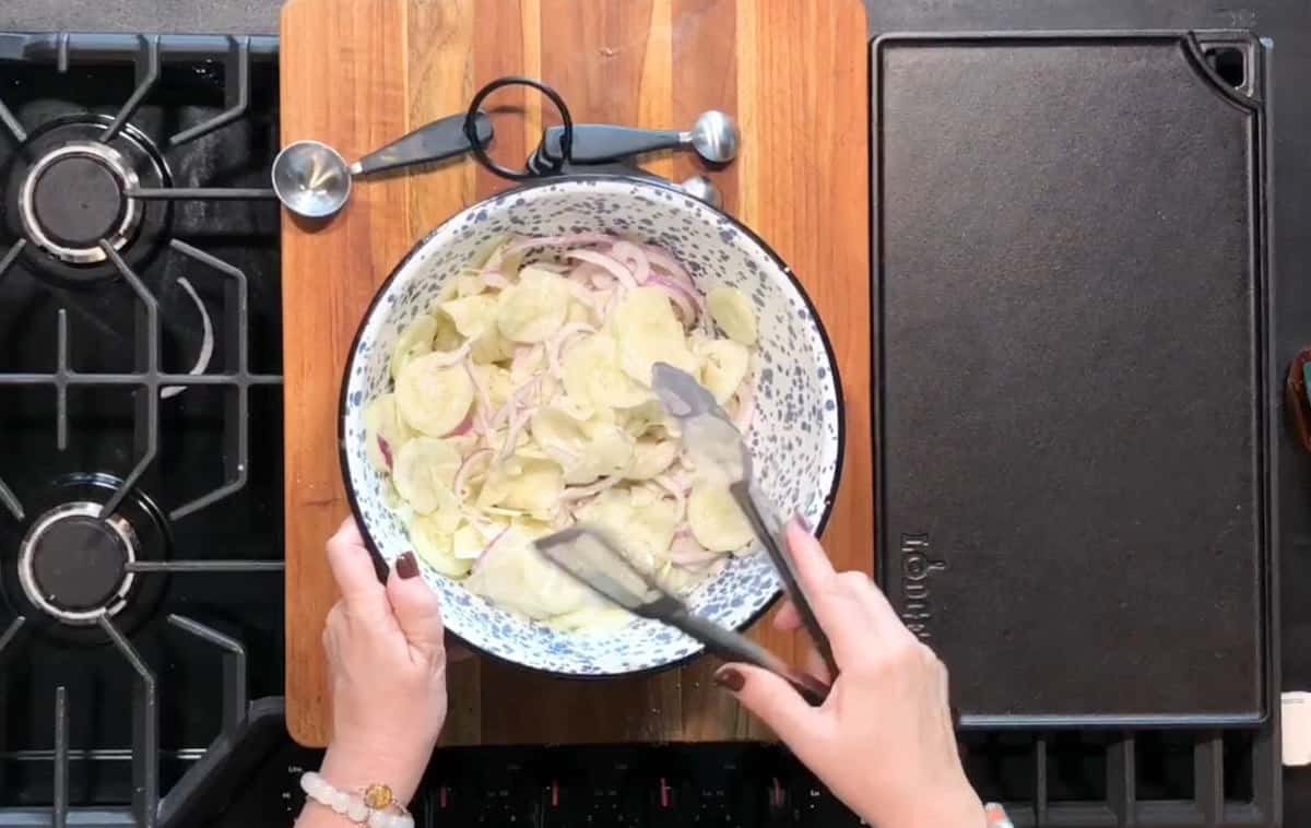 Hands are using tongs to mix sliced potatoes and onions in a large, speckled bowl on a wooden cutting board. Measuring spoons are nearby. The scene is set on a stove with a griddle on the right.