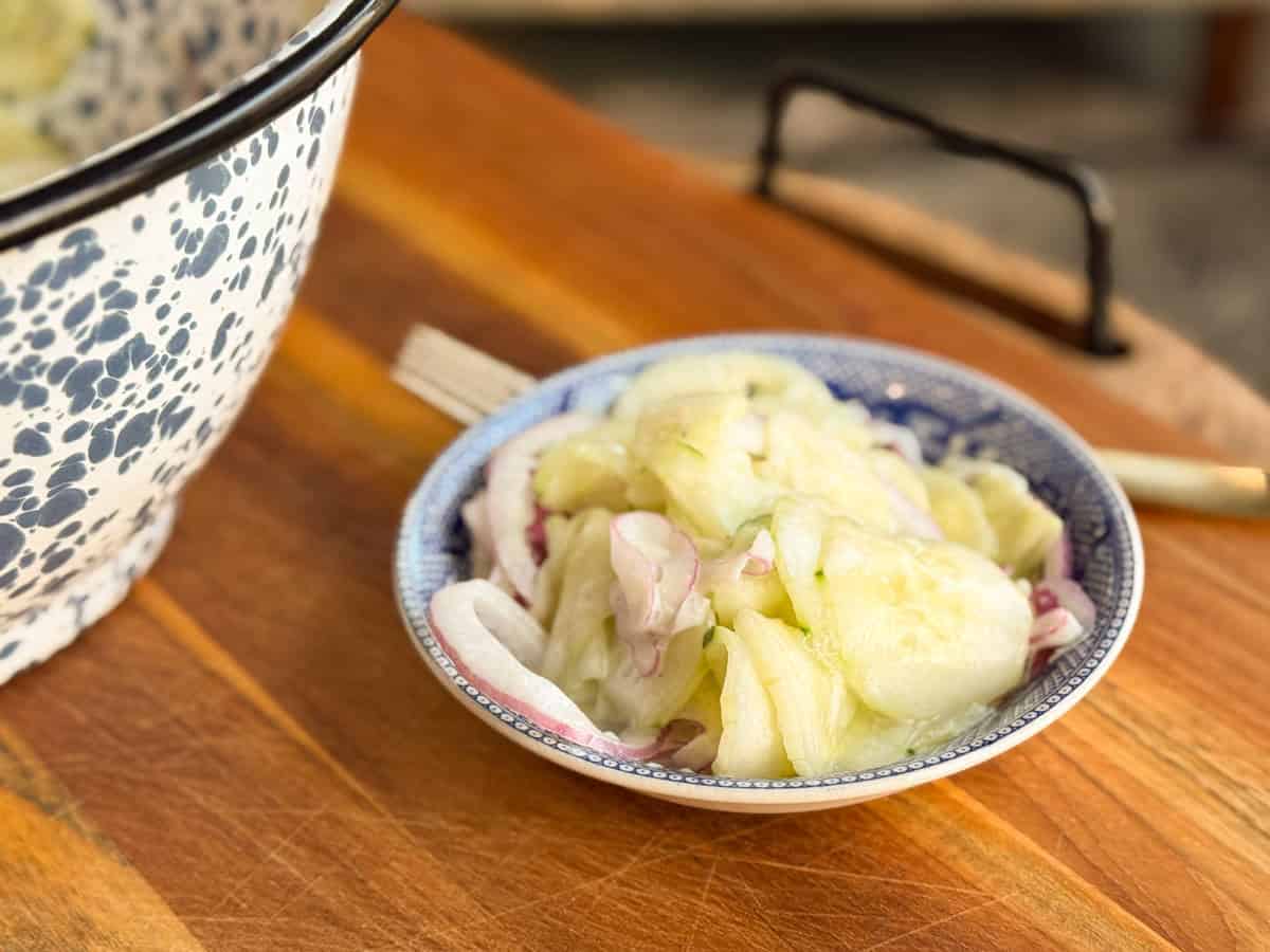 A small blue and white bowl sits on a wooden surface, filled with sliced cucumbers and onions. A large patterned basin is partially visible in the background. A fork lies next to the bowl.