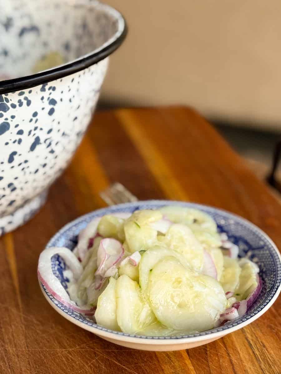 A close-up of a small dish containing sliced cucumbers and red onions, placed on a wooden surface. Behind it is a large bowl with a speckled design. A fork is partially visible near the dish.