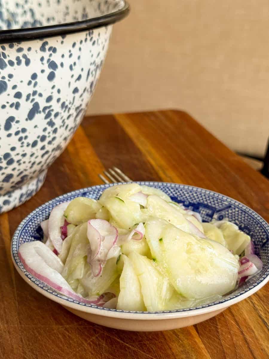 A bowl of cucumber salad with sliced cucumbers and red onions in a creamy dressing is on a wooden table. A large speckled mixing bowl and a fork are in the background.
