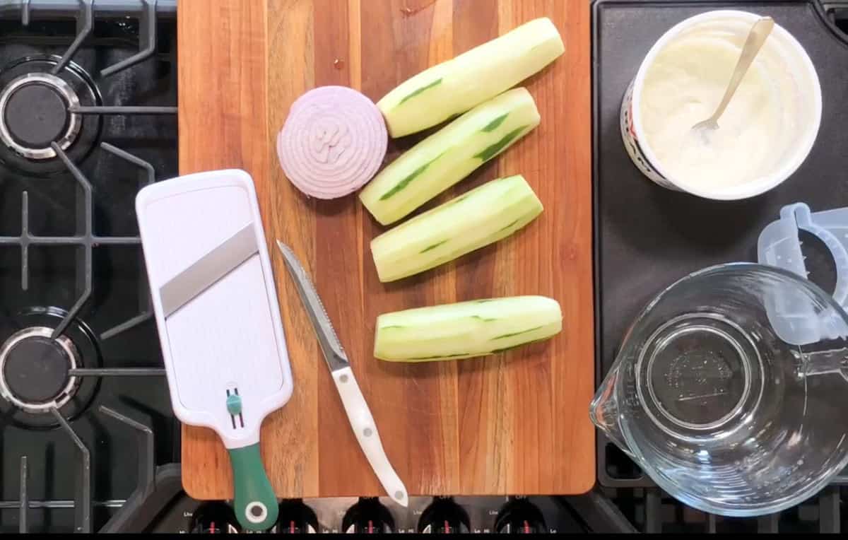 A kitchen countertop with a wooden cutting board. On the board are a mandoline slicer, a knife, a peeled onion, and three peeled cucumbers. To the side, there is a container with white sauce and a clear measuring jug. Stove burners are visible.
