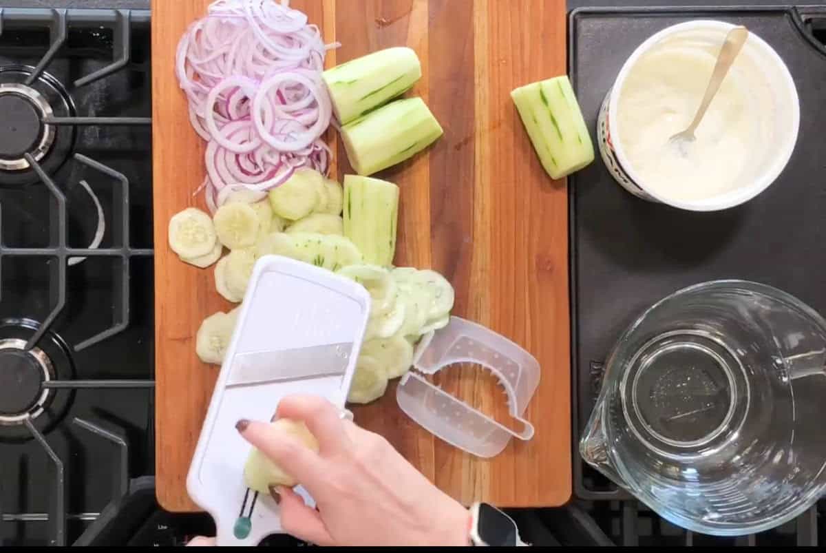Hand slicing cucumbers using a mandoline on a wooden cutting board. Sliced red onions, a bowl of cream or dressing, and a clear pitcher are nearby on a black countertop with a stove on the left.