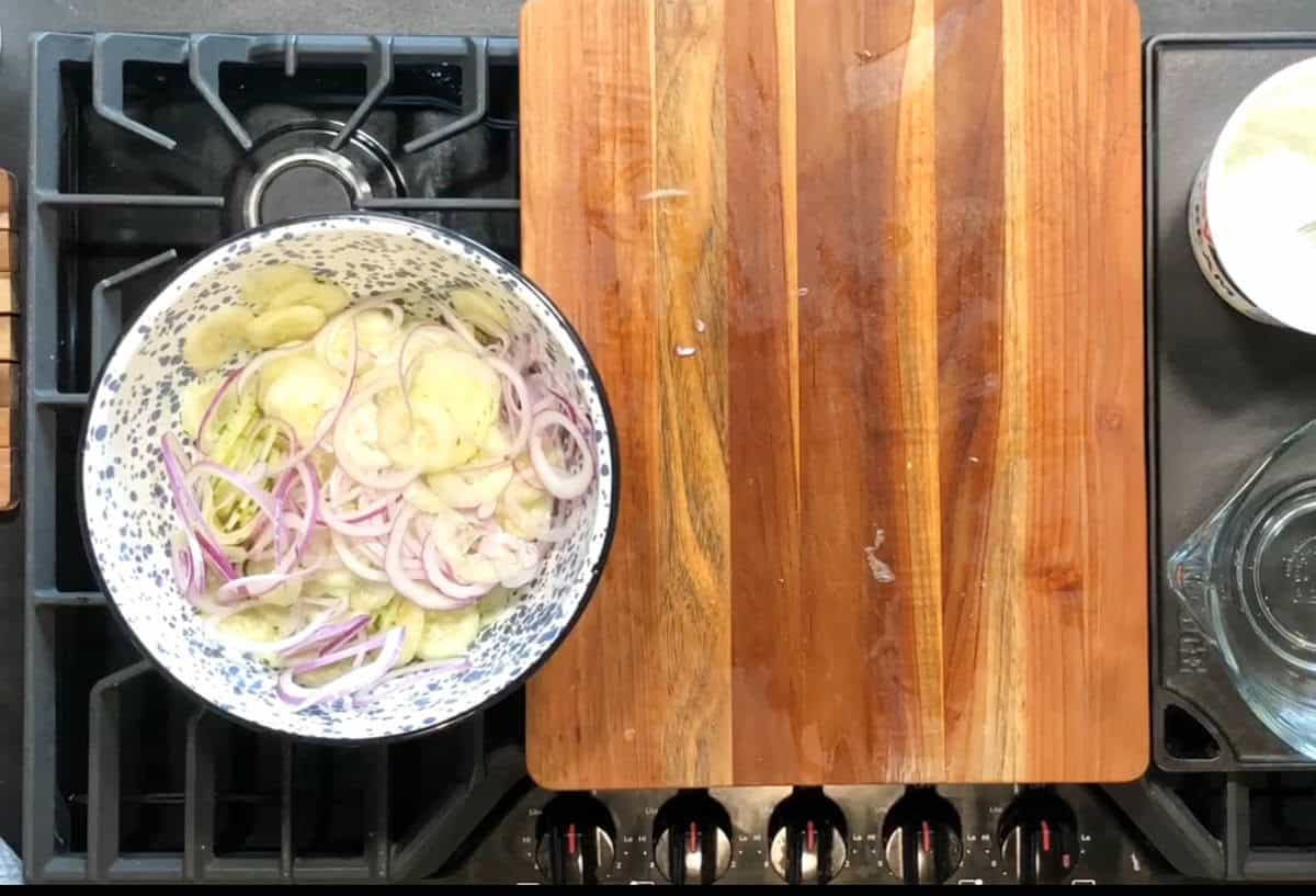 A kitchen stove with a wooden cutting board in the center. To the left, there's a bowl containing sliced onions and cucumbers. A measuring cup is partially visible on the right side of the stove.