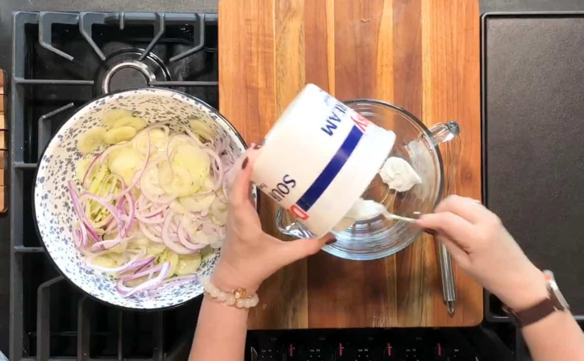 A person is scooping sour cream from a container into a glass bowl on a wooden cutting board. Next to it, there is a large bowl filled with sliced potatoes and onions. The setup is on a kitchen stove.