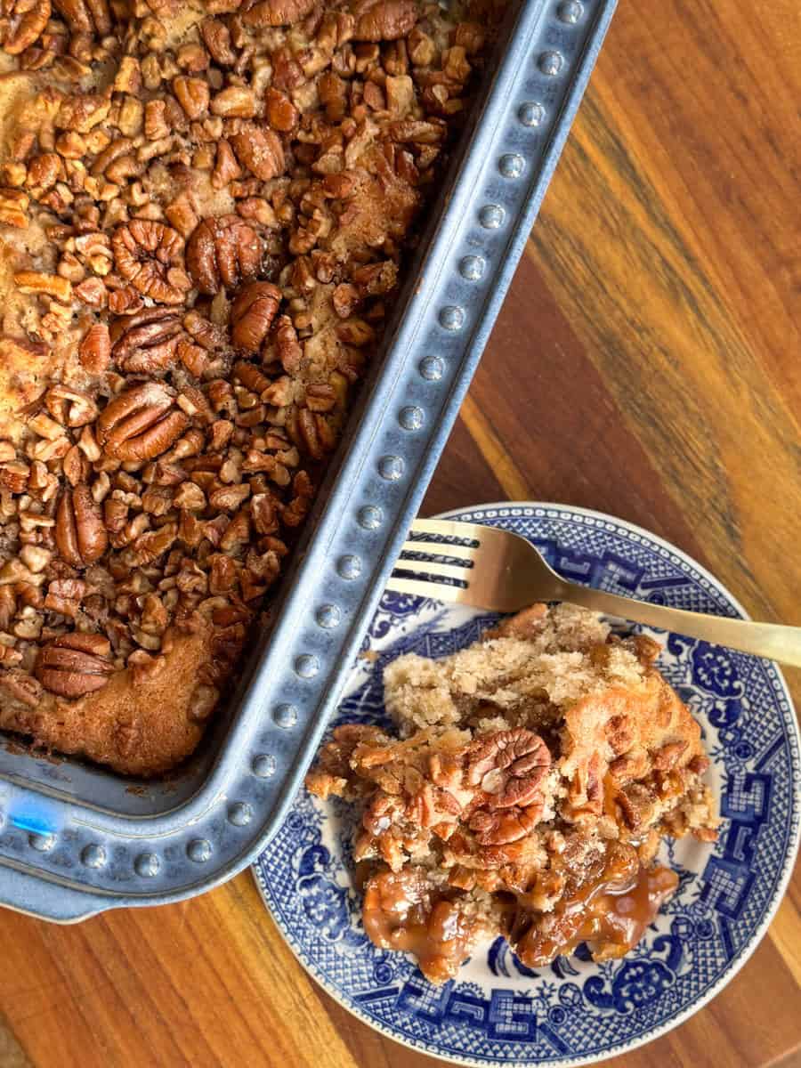 Baking dish filled with pecan-topped dessert on a wooden table. A piece is served on a blue-patterned plate with a fork, showing a caramel-like filling under the topping.