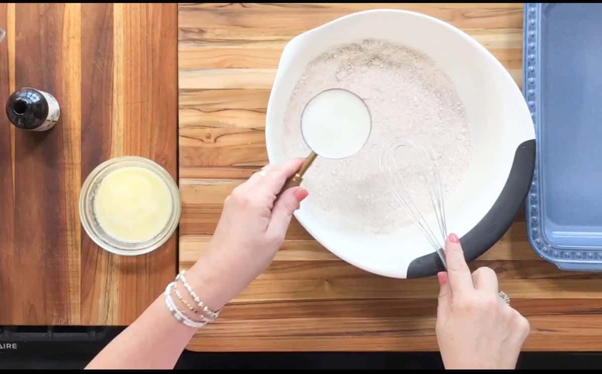 A person is holding a measuring cup of milk over a mixing bowl containing dry ingredients. An empty glass dish, a small bowl of melted butter, and vanilla extract are on the wooden countertop. A whisk is resting in the mixing bowl.
