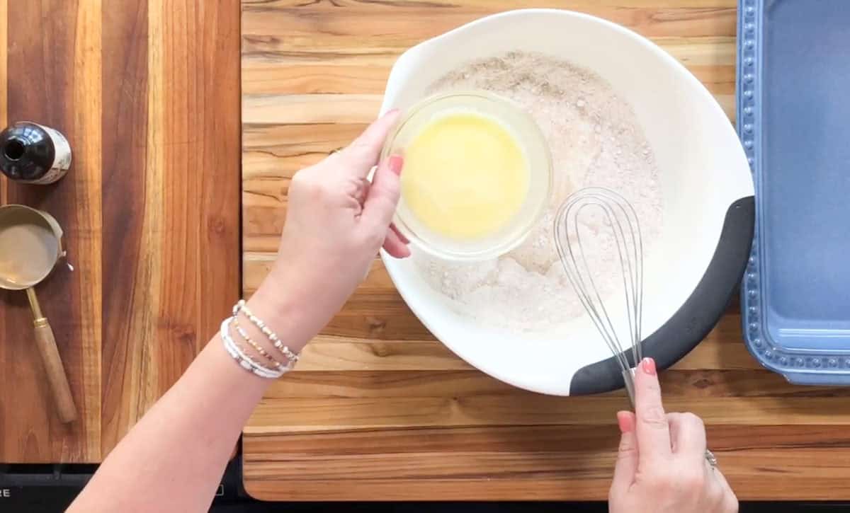 Hands are shown adding a small bowl of melted butter to a larger mixing bowl containing dry ingredients. A whisk is in the large bowl. A blue baking dish, vanilla extract, measuring cup, and spoon are on a wooden surface nearby.