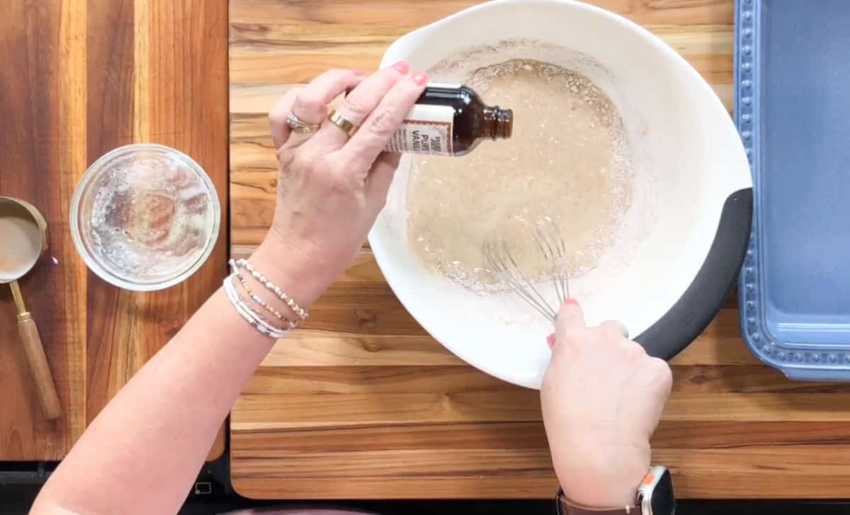 Hands are mixing ingredients in a white bowl with a whisk on a wooden surface. A bottle of brown liquid is being poured in. Nearby are a small glass bowl with residue and a silver measuring cup. A blue rectangular dish is on the right side.