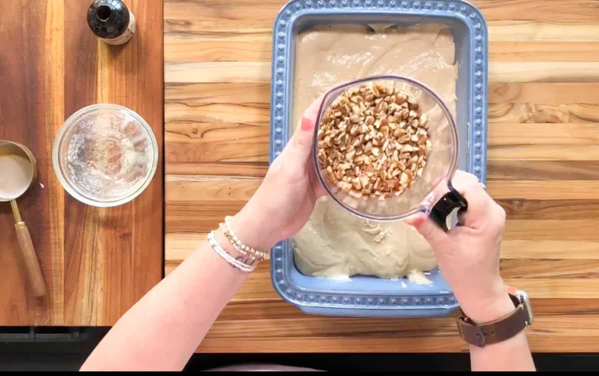 A person is holding a cup filled with chopped nuts over a baking dish containing batter. The baking dish is on a wooden surface. Nearby are a glass bowl with residue, a wooden spoon, and a bottle with a dark liquid.