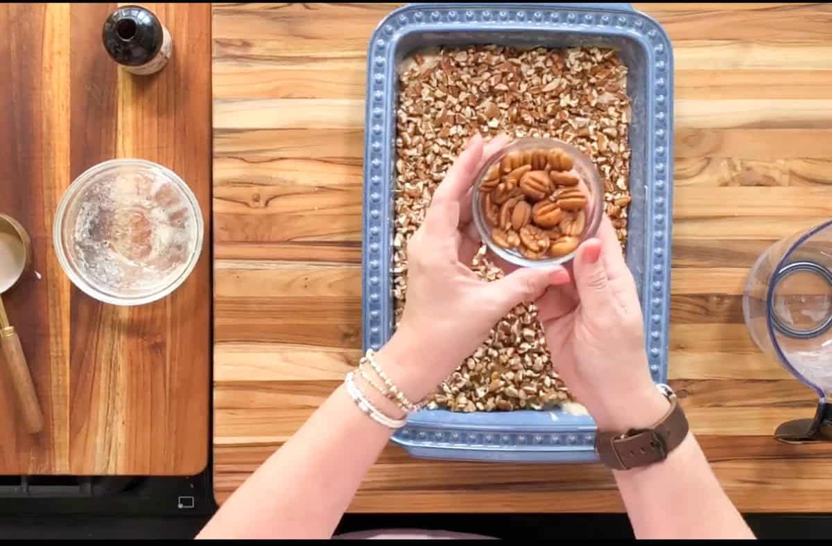 A person holds a small bowl of pecans over a blue baking dish filled with chopped nuts. The scene is on a wooden surface with a glass bowl, a bottle, and a spoon nearby.