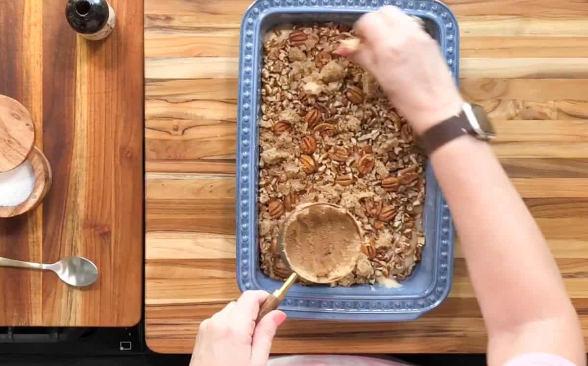 Person spreading a mixture in a blue baking dish on a wooden surface. The dish contains pecans and other ingredients. Nearby are a small bottle, spoon, and salt container. The person is wearing a watch on their left wrist.