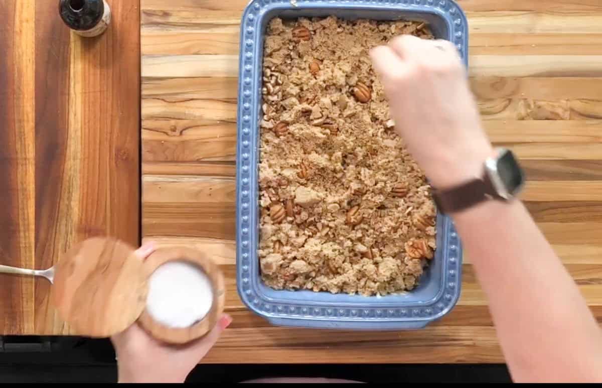 Hands are sprinkling an ingredient from a wooden container onto a dish with a crumbly topping in a blue baking dish. The dish is on a wooden countertop, and a small bottle is visible nearby. The person is wearing a watch.