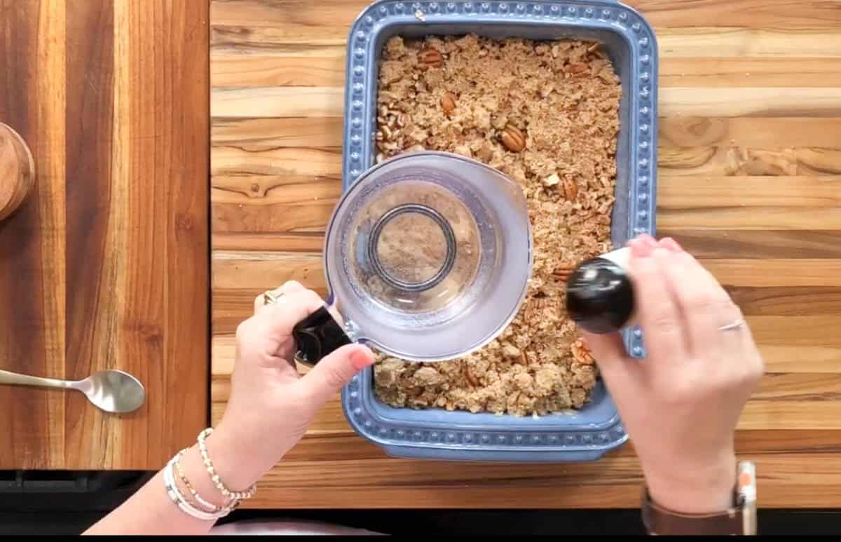 A person is about to pour a liquid from a measuring cup over a blue baking dish filled with a crumbly mixture, possibly granola. The scene is set on a wooden surface, and a spoon is visible to the side.