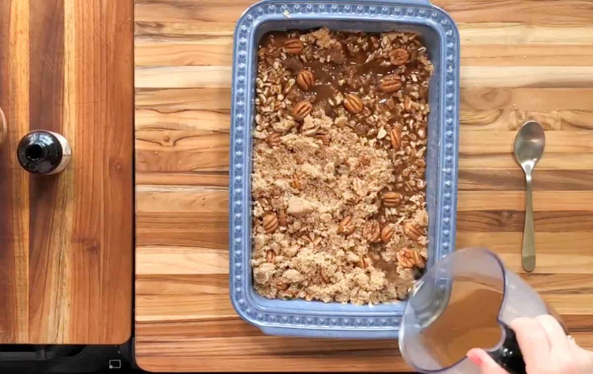 The image shows a blue rectangular baking dish on a wooden surface, containing a partially unbaked dessert with pecans and crumble topping. A hand is pouring liquid from a blender into the dish. A spoon and a bottle are nearby.