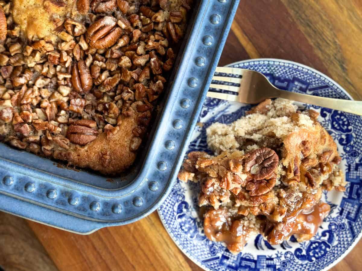 A blue baking dish with pecan-topped dessert next to a blue and white patterned plate holding a serving of the dessert. A fork rests on the plate. The wooden table provides a warm background.