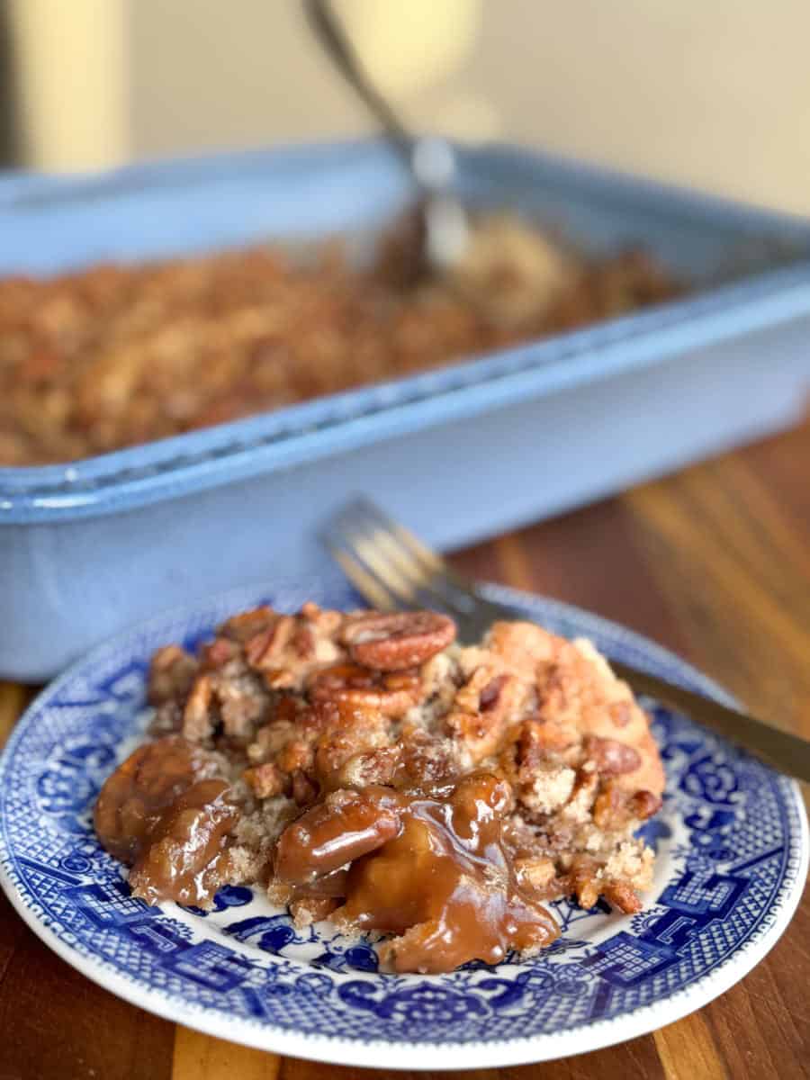 A slice of pecan-topped dessert with gooey filling on a blue and white plate. A fork is placed beside it. In the background, a blue baking dish holds the remaining dessert with serving spoon visible.