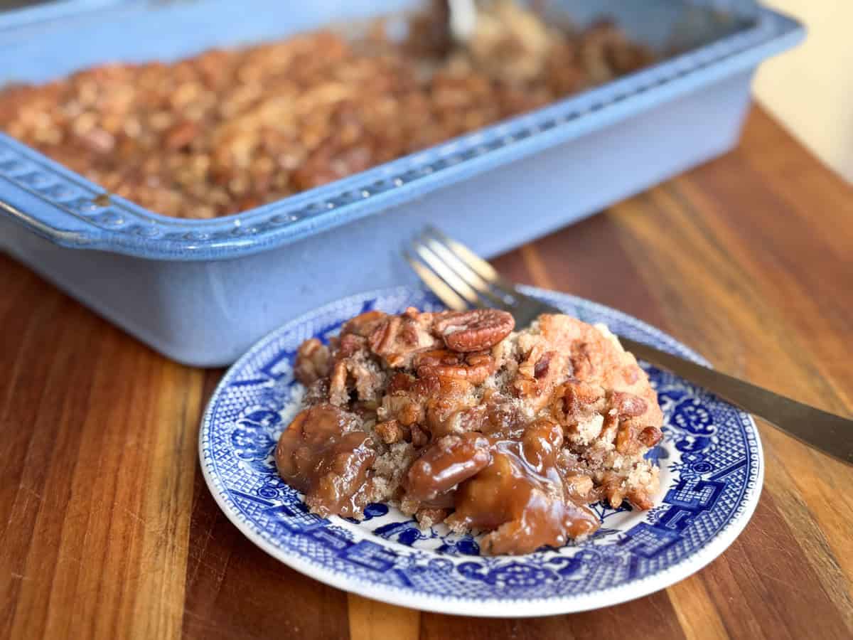 A dessert with a golden-brown topping is served on a blue and white patterned plate, placed on a wooden surface. The dish features pecans and a gooey, caramelized filling. A fork and the larger baking dish with the dessert are in the background.