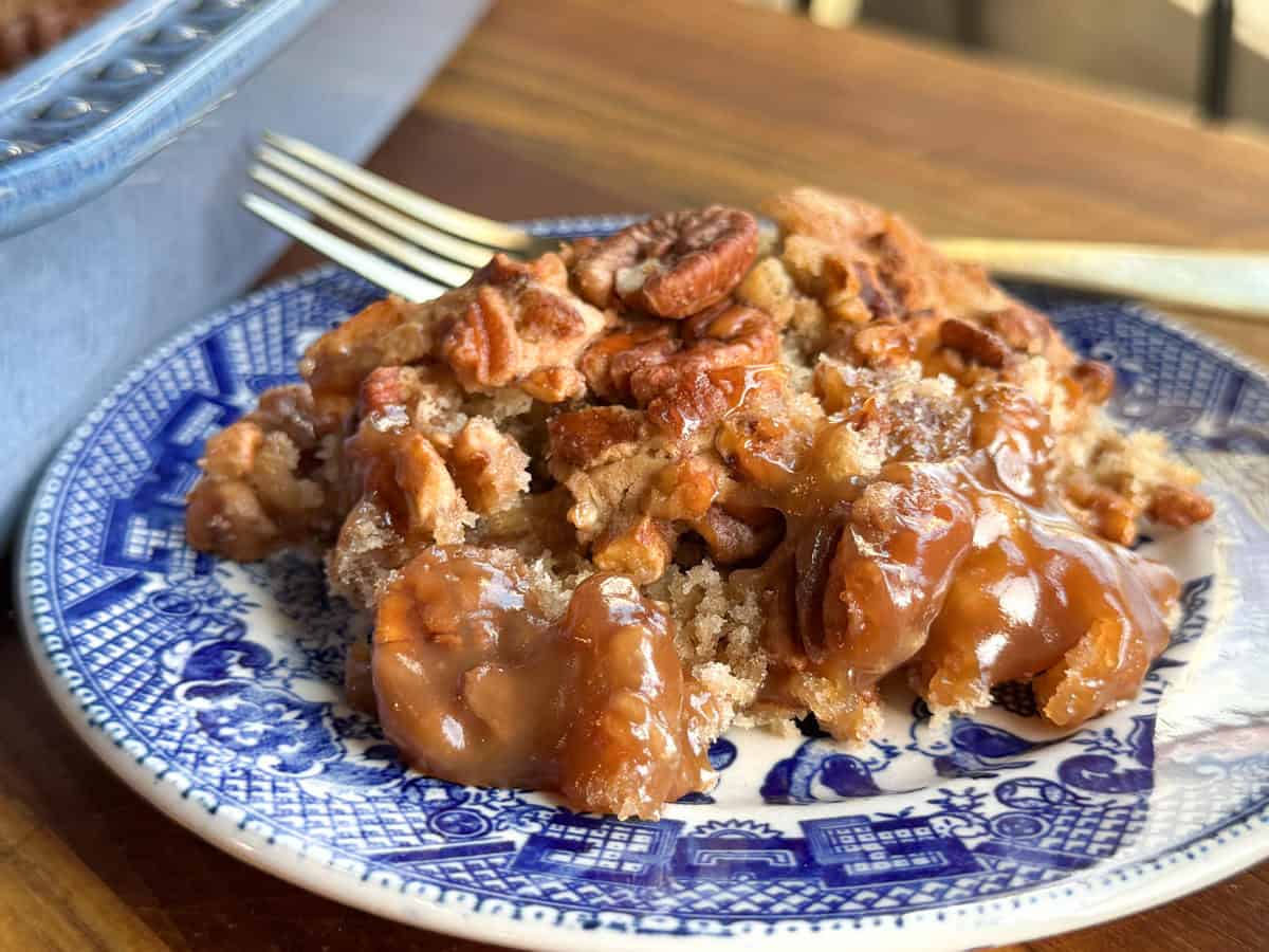 A slice of pecan dessert with caramel sauce on a blue and white patterned plate, placed on a wooden table. A fork is visible in the background.