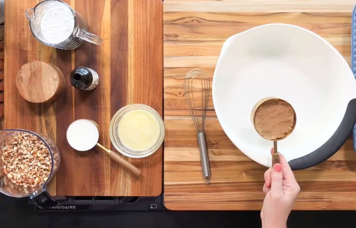 A person holds a measuring cup over a large empty bowl. On the wooden countertop are chopped nuts, flour, vanilla extract, sugar, melted butter, and a whisk.