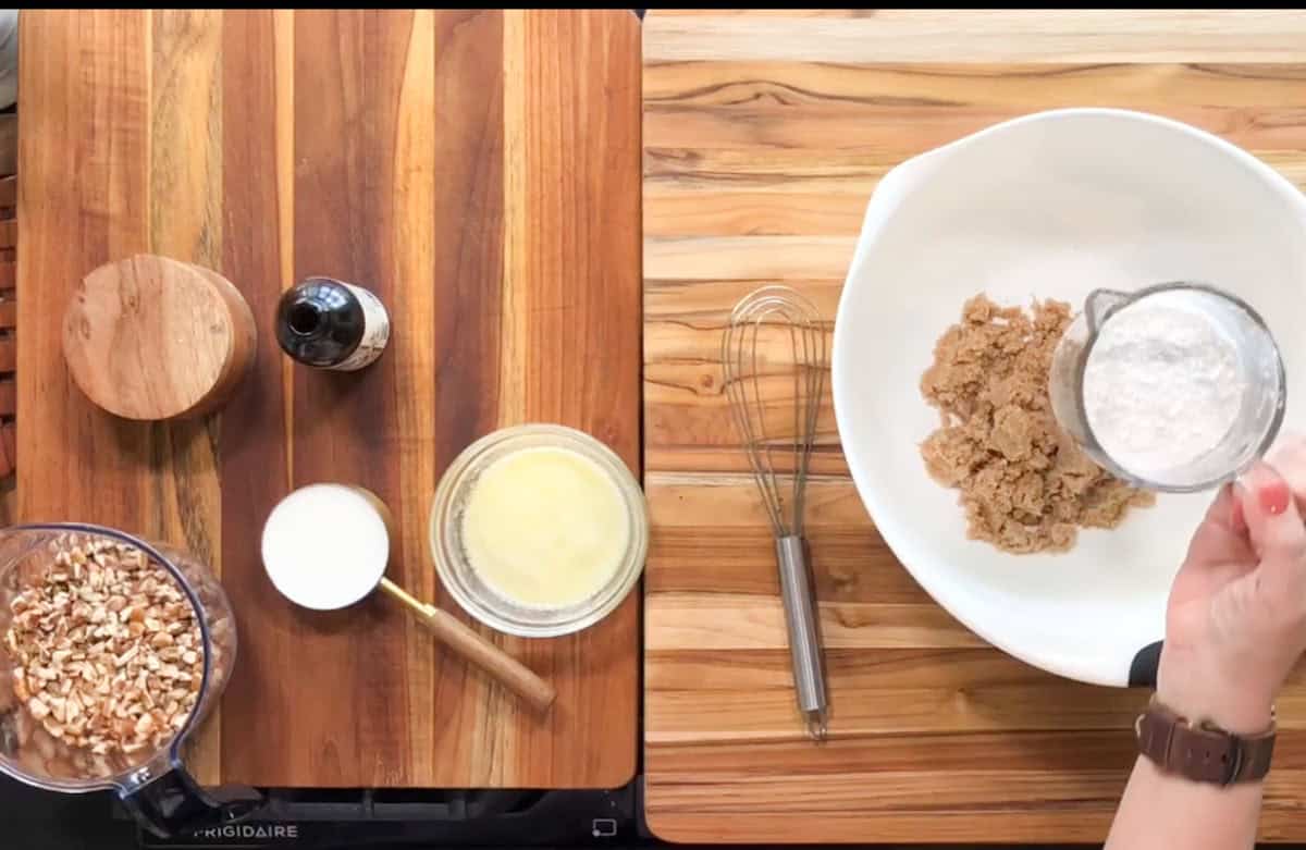 A kitchen countertop with ingredients for baking. On the left, a food processor with chopped nuts, a wooden container, a bottle, and a bowl of milk. On the right, a bowl with brown sugar being mixed with flour, next to a whisk.