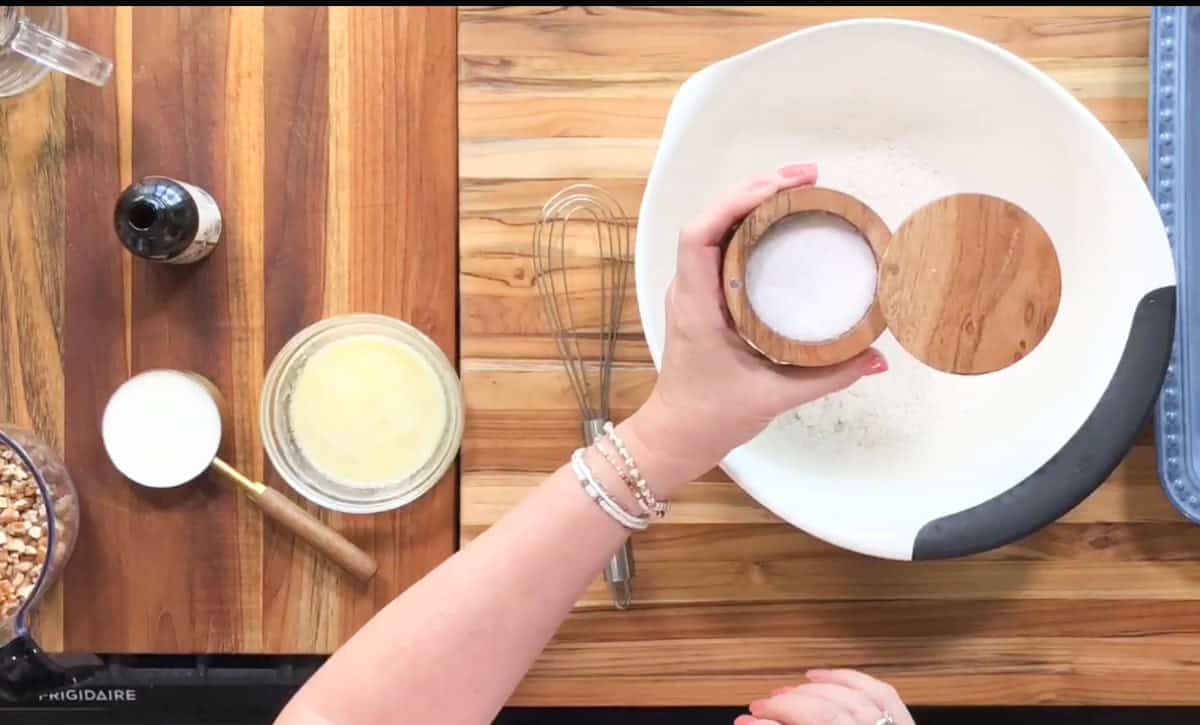 A hand is holding a small wooden container over a white mixing bowl with flour. Nearby are a whisk, a bowl with melted butter, a measuring cup with sugar, vanilla extract, and chopped nuts, all on a wooden surface.