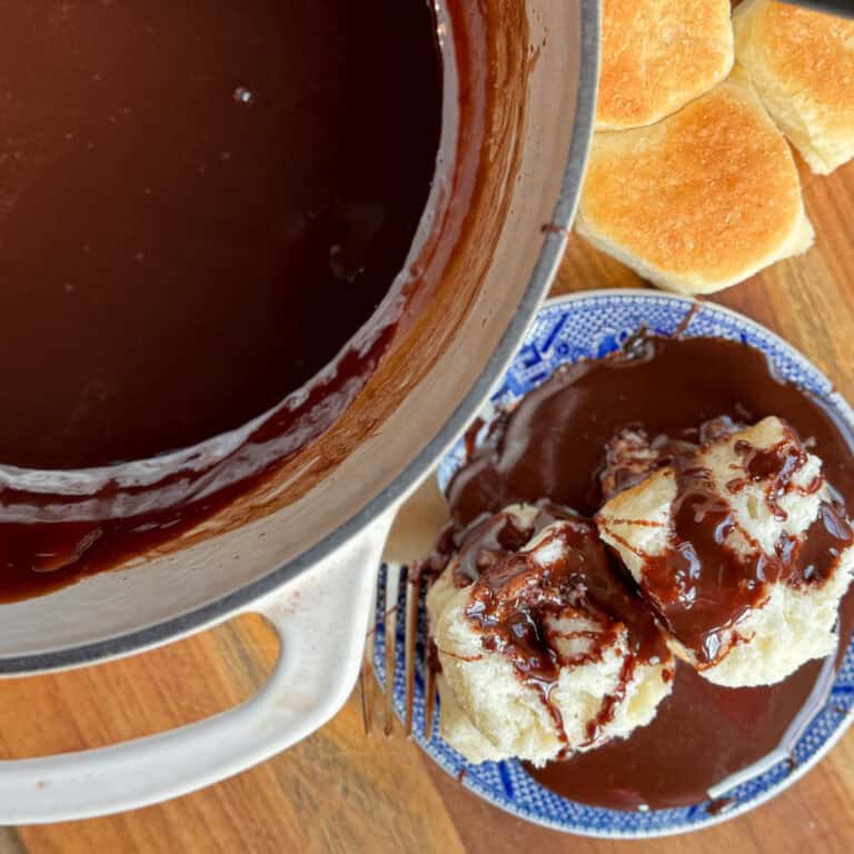 A pot of rich chocolate gravy sits on a wooden surface beside a blue-patterned plate with two biscuits covered in gravy. Several plain biscuits are arranged nearby. A fork rests near the plate.